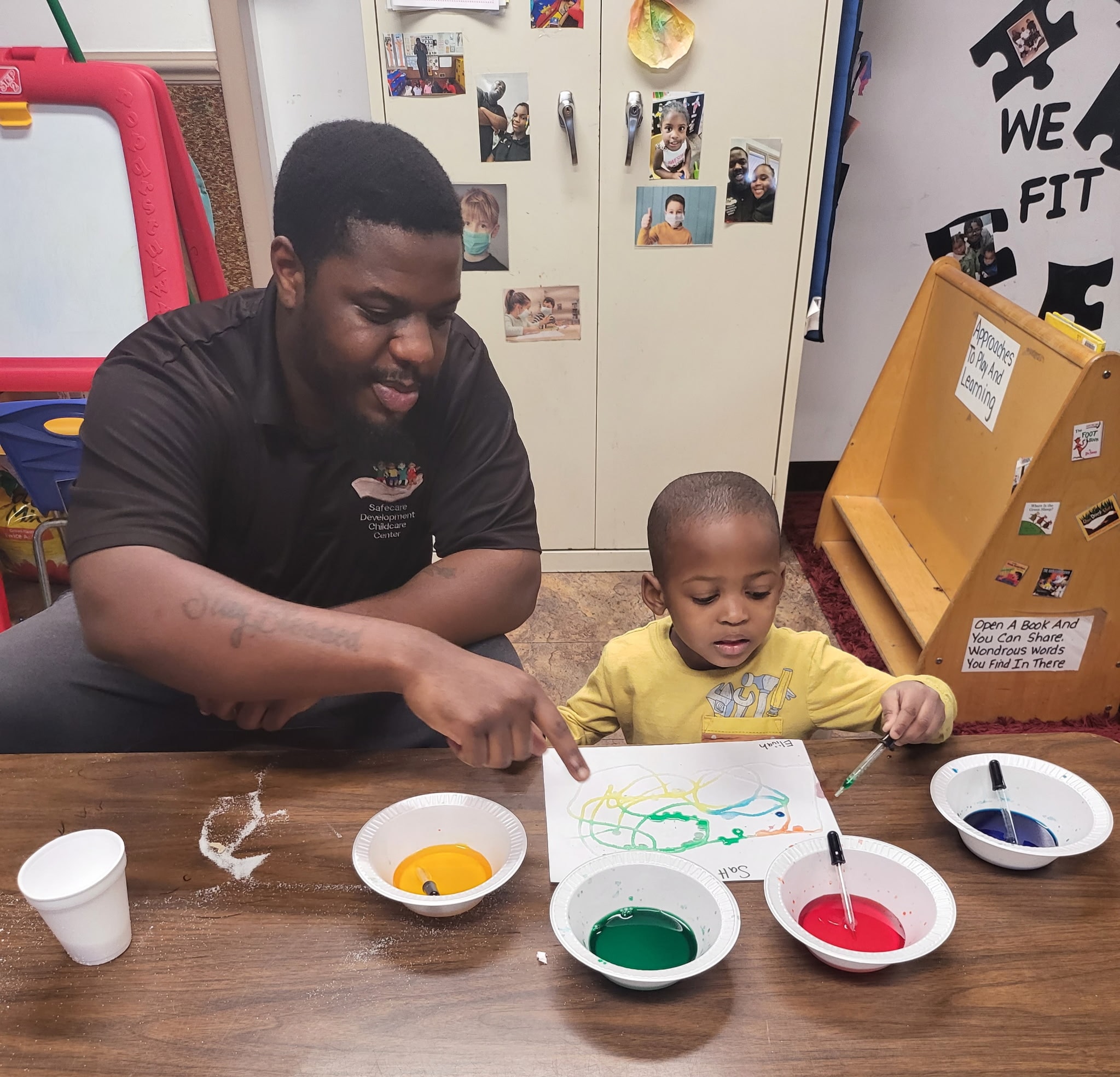 Safecare staff member guiding a toddler through a colorful painting activity at the classroom table