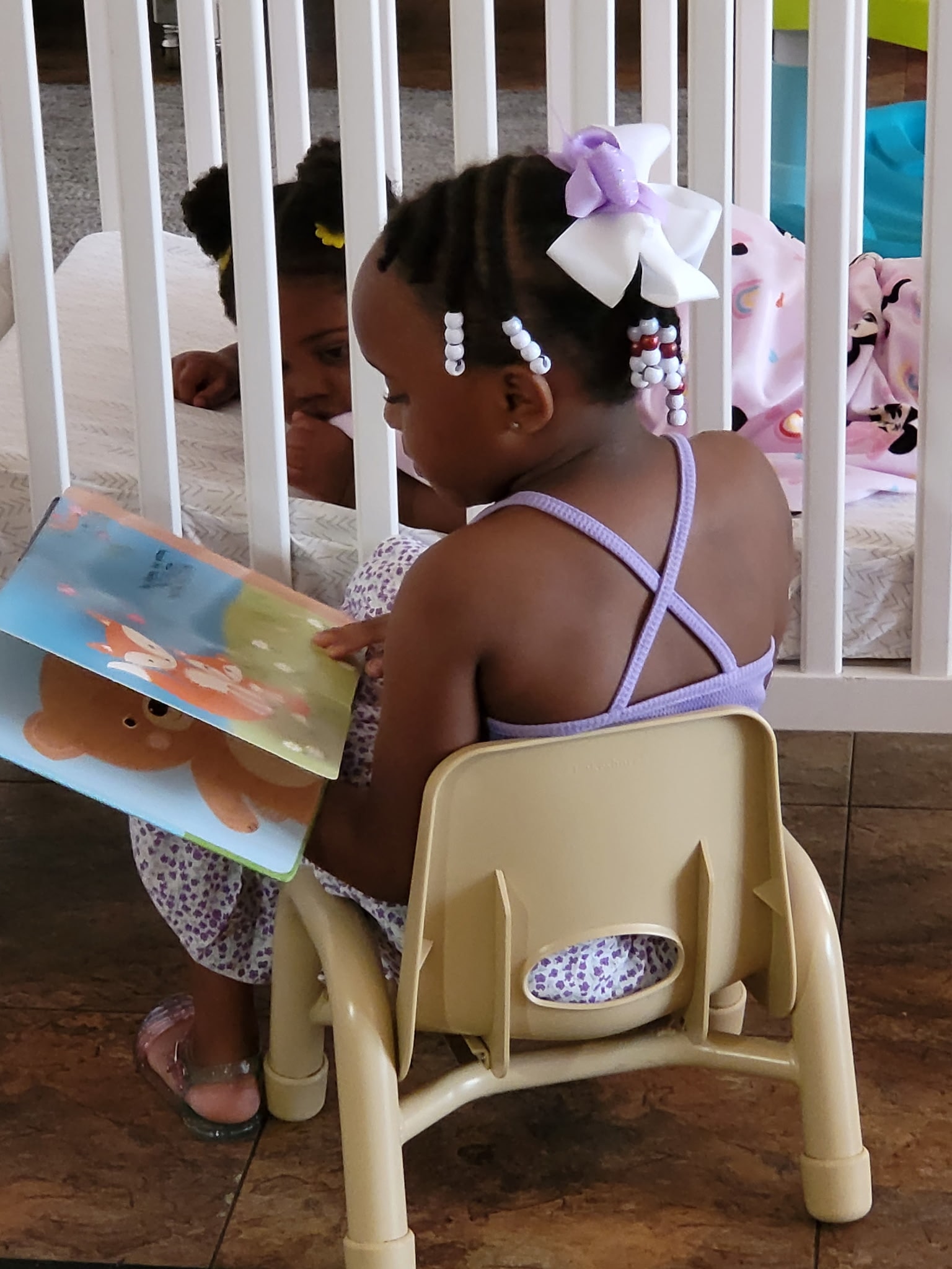 Toddler girl sitting in a chair reading a picture book next to a crib in the classroom